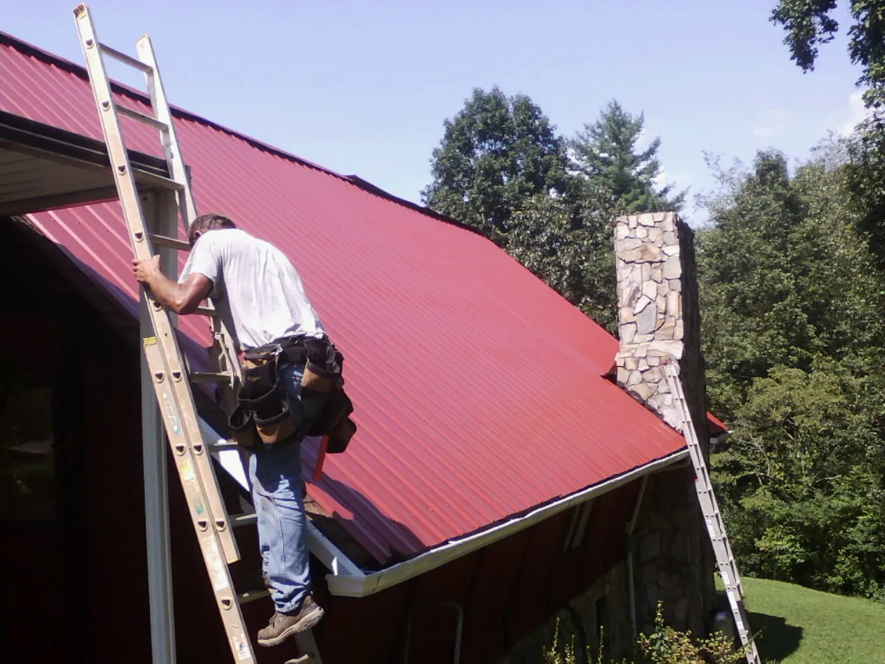 Roofer on a ladder beside a red metal roof and stone chimney — Soffit & Fascia Repair by Roofing Brothers in Binghamton